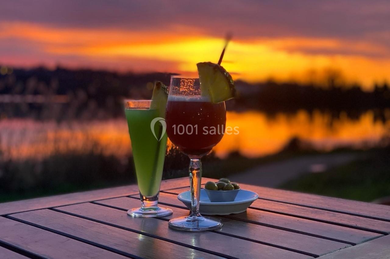 Deux cocktails colorés et une assiette d'olives sur une table en bois, avec un coucher de soleil en arrière-plan.