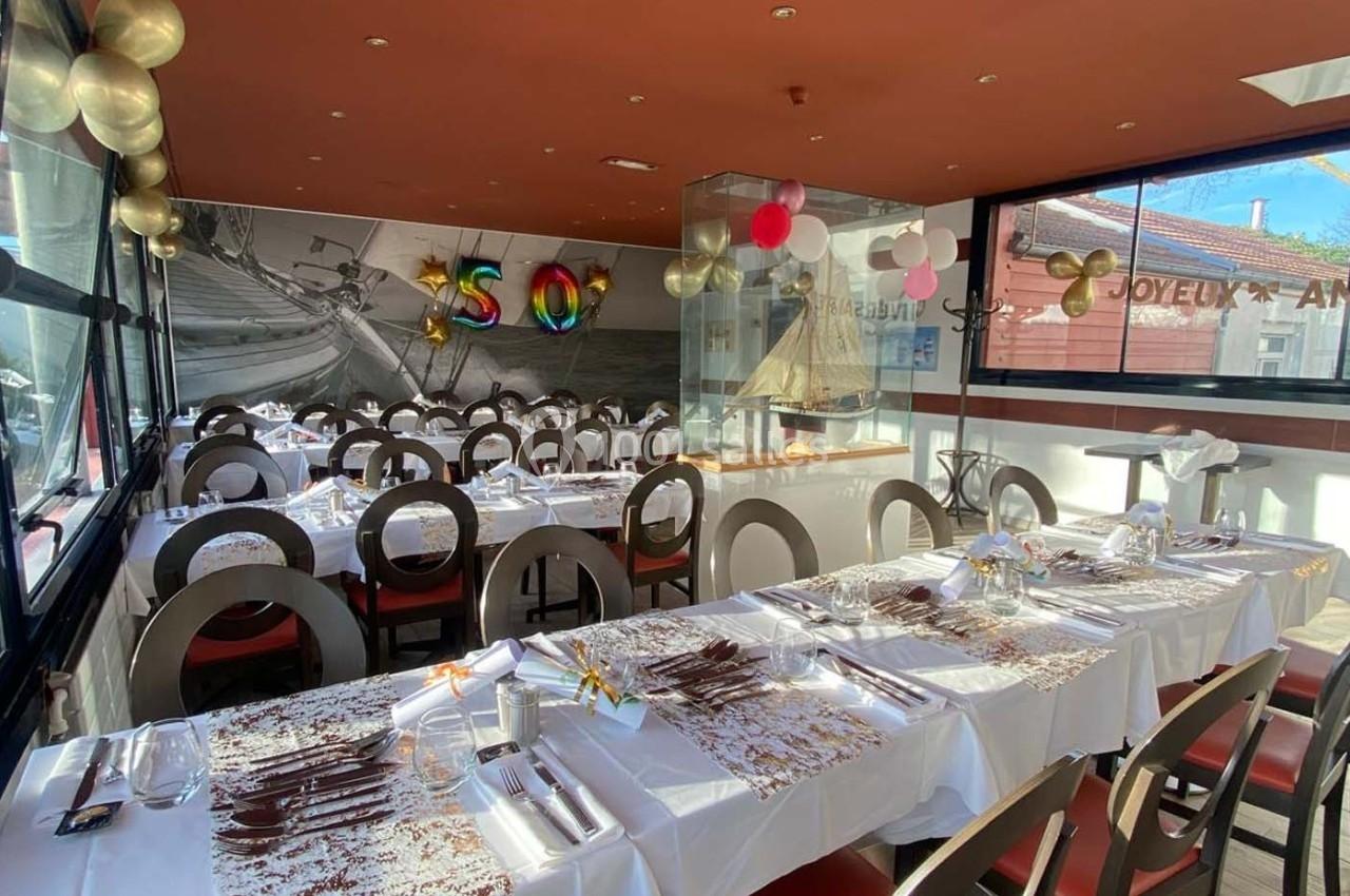 Salle de restaurant décorée pour une fête, avec tables dressées, ballons colorés et maquette de bateau en arrière-plan.