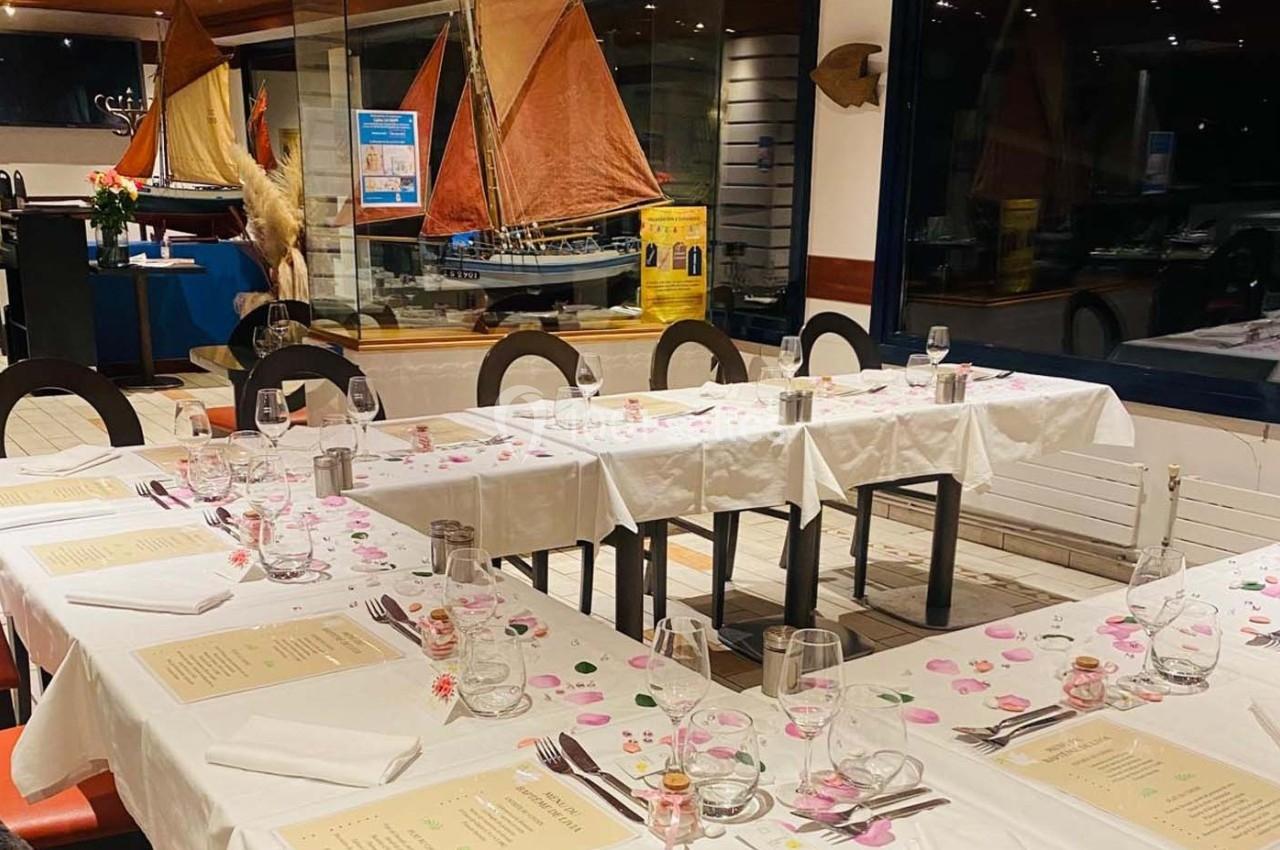 Salle de restaurant avec tables dressées, nappes blanches, menus et décoration maritime en arrière-plan.