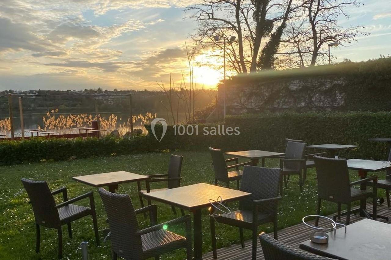 Terrasse extérieure avec des tables et des chaises en rotin, vue sur un lac au coucher du soleil.