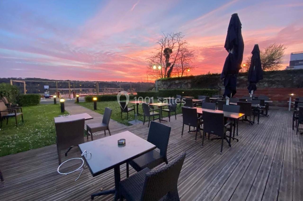 Terrasse en bois avec tables et chaises vides, vue sur un coucher de soleil coloré et un paysage dégagé.