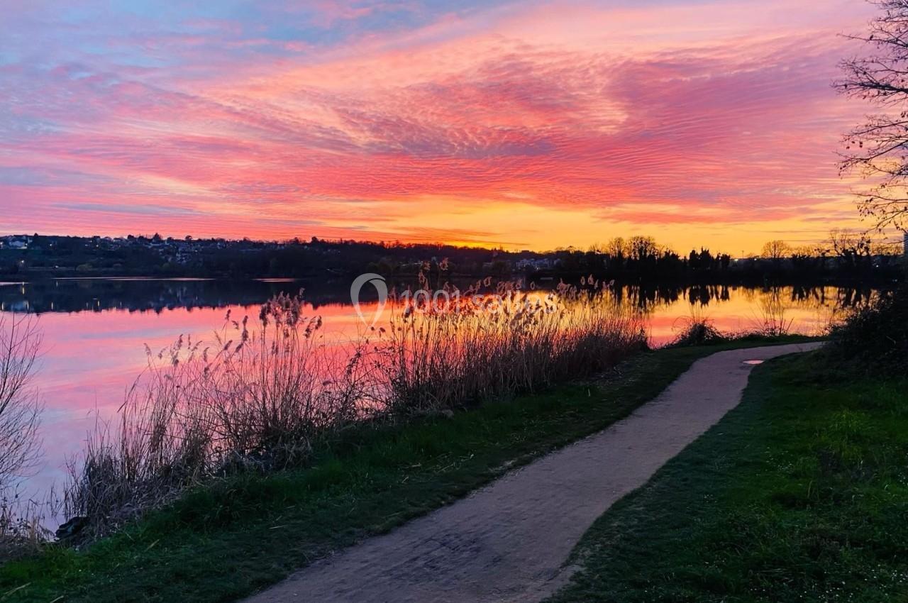 Chemin bordé d'herbes longeant un lac au coucher du soleil, avec un ciel aux teintes roses et orangées.