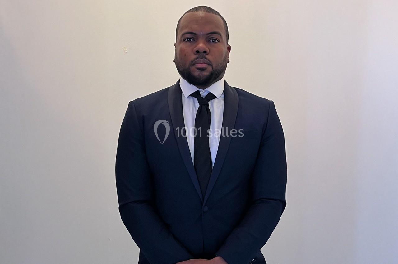 Homme en costume sombre et cravate noire, debout devant un mur blanc.
