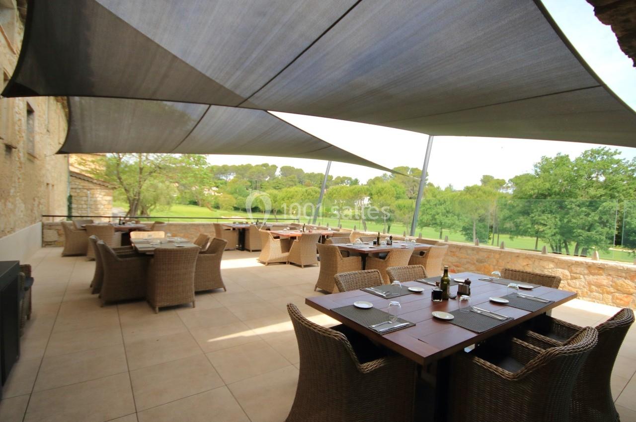 Terrasse extérieure avec tables en bois, chaises en rotin et voiles d'ombrage, donnant sur un paysage verdoyant.