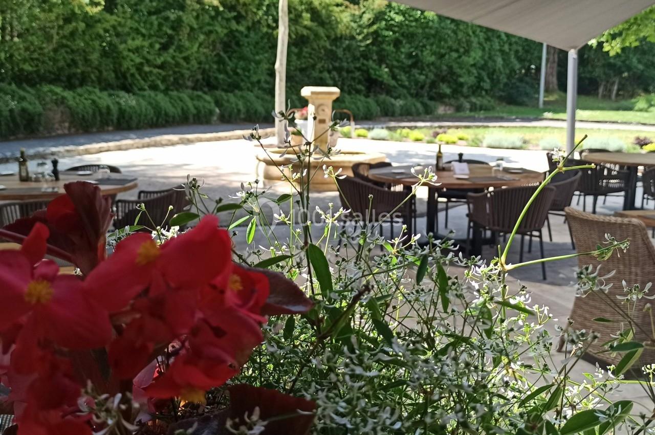 Terrasse extérieure avec tables en rotin, vue sur un jardin verdoyant et des fleurs rouges au premier plan.