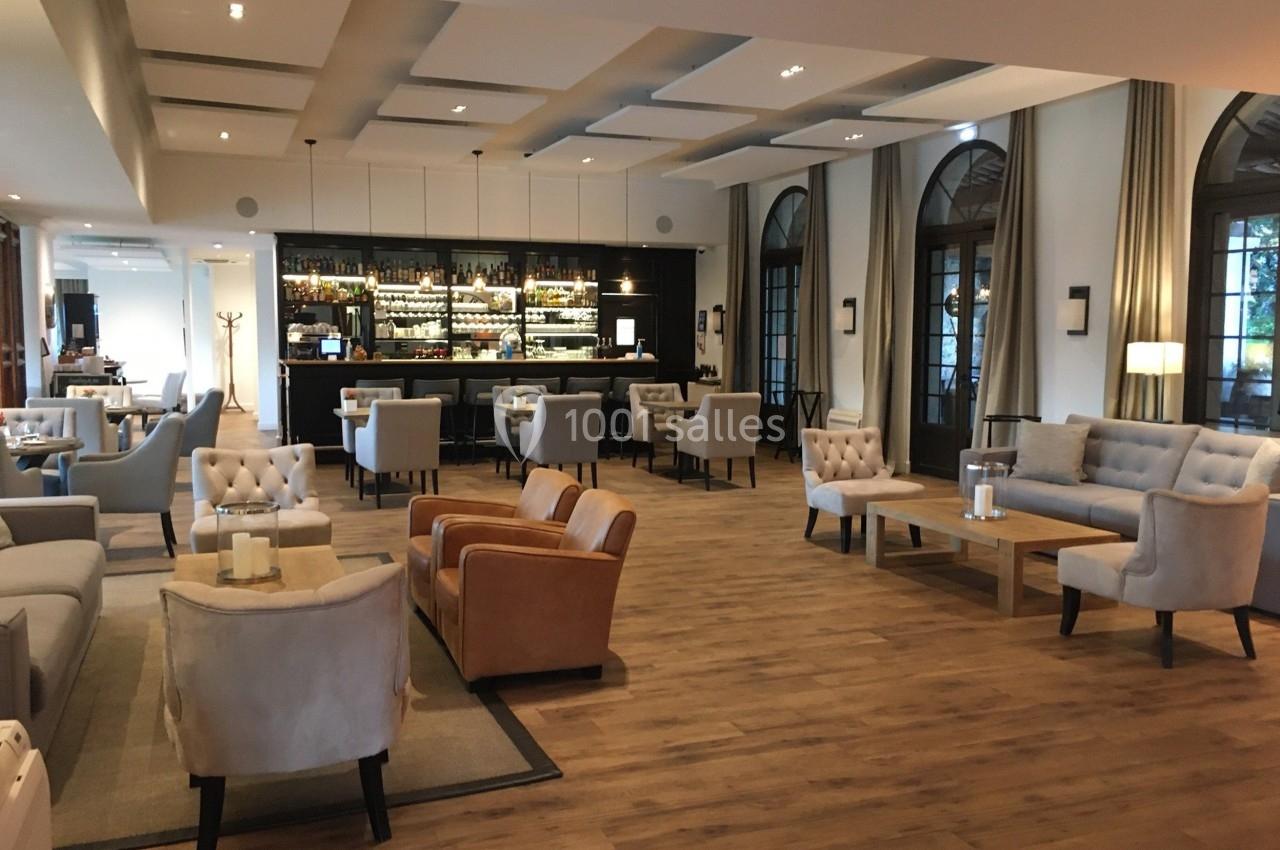 Salon moderne avec parquet, fauteuils confortables, tables basses et un bar éclairé en arrière-plan.