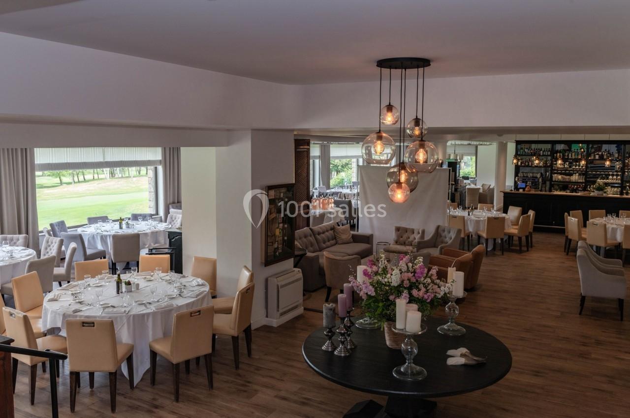 Salle de restaurant élégante avec tables dressées, grandes fenêtres donnant sur un espace vert et un bar en arrière-plan.