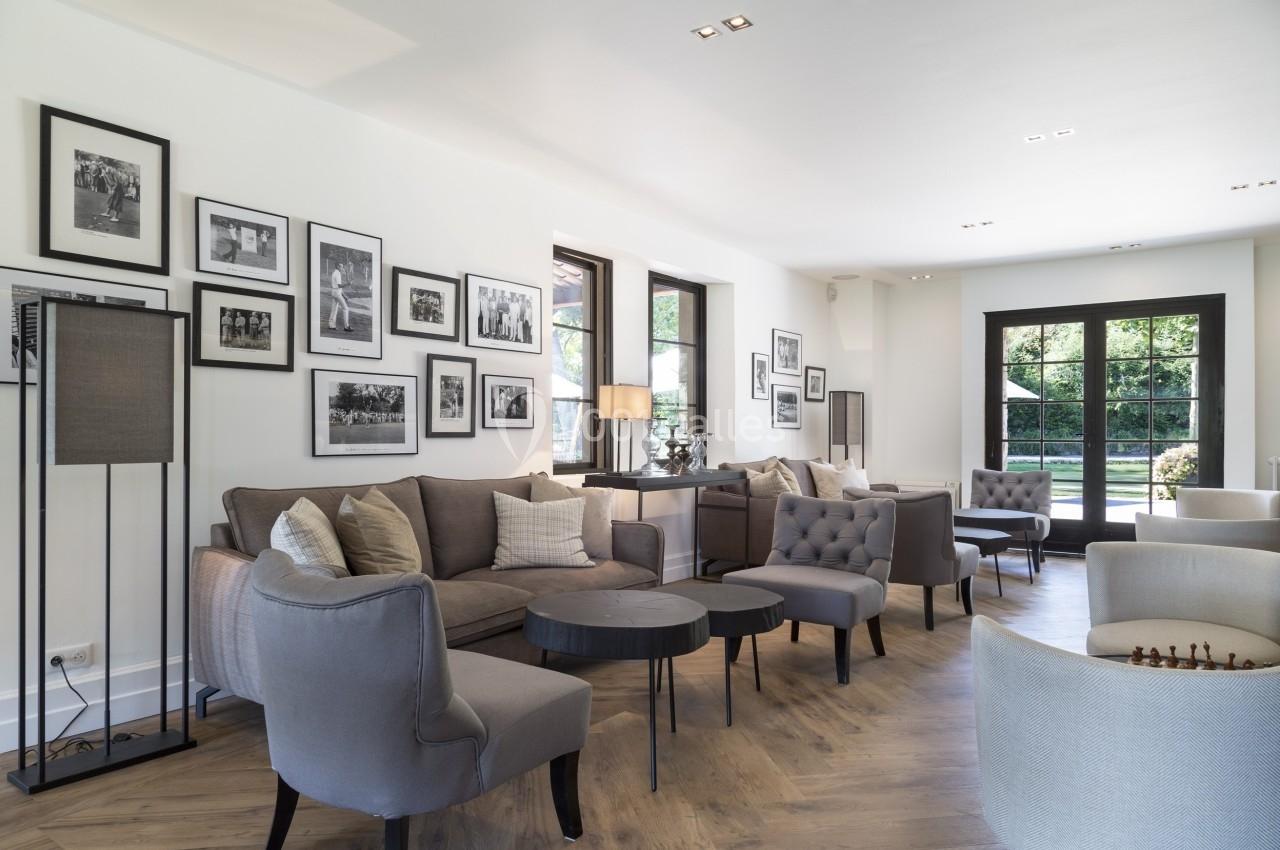 Salon lumineux avec canapés gris, tables basses rondes, murs décorés de cadres et grandes fenêtres donnant sur un jardin.