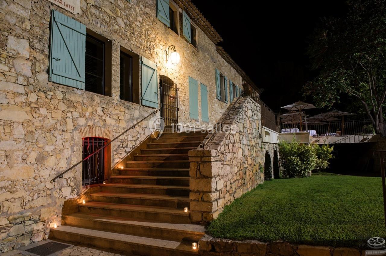 Escalier en pierre éclairé menant à une maison en pierre avec volets bleus, entourée de pelouse et d'arbres.