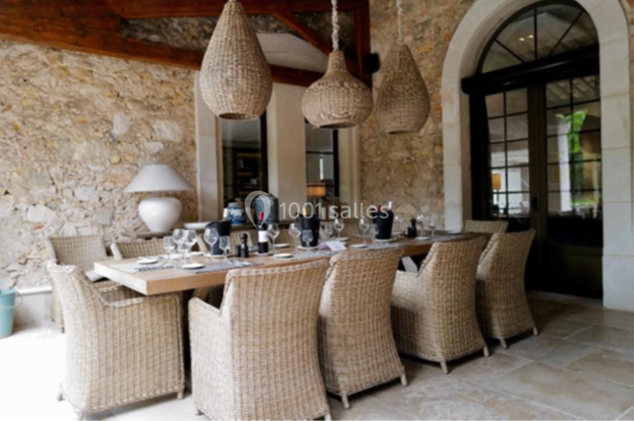 Salle à manger extérieure avec table en bois, chaises en rotin, suspensions tressées et murs en pierre apparente.