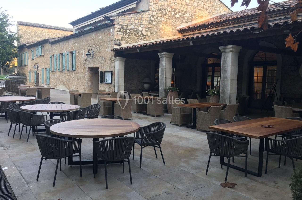 Terrasse d'un restaurant avec des tables en bois et des chaises en rotin, devant un bâtiment en pierre.