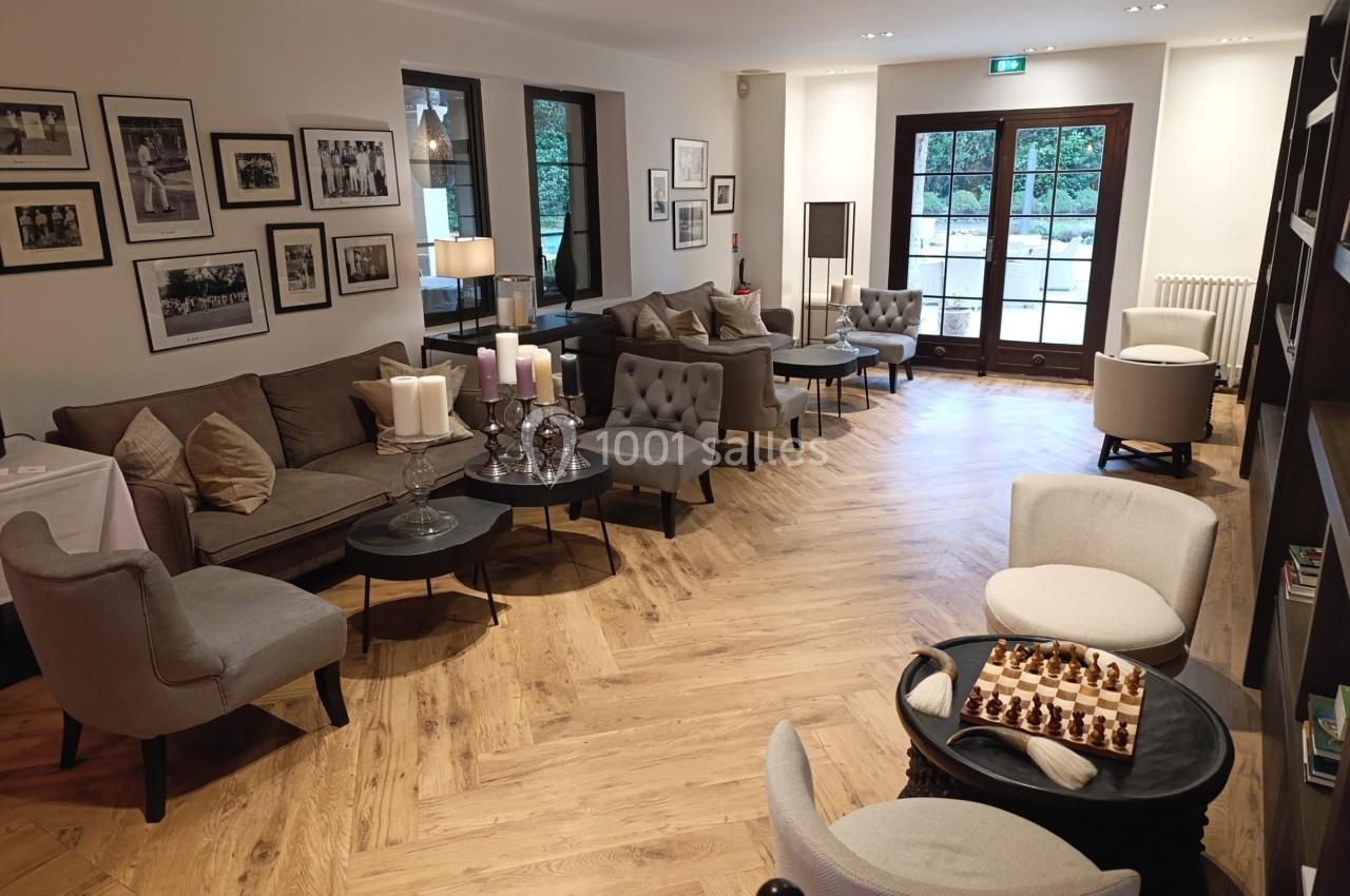 Salon lumineux avec parquet en bois, canapés, fauteuils, tables basses, décorations murales et échiquier sur une table.