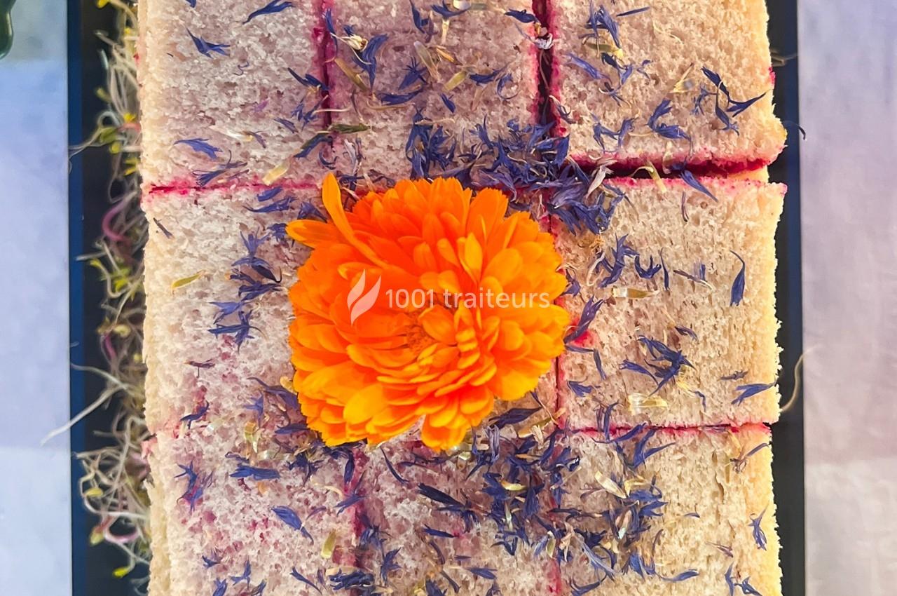 Gâteau rectangulaire décoré de fleurs séchées violettes et d'une fleur orange centrale, présenté sur un plateau noir.