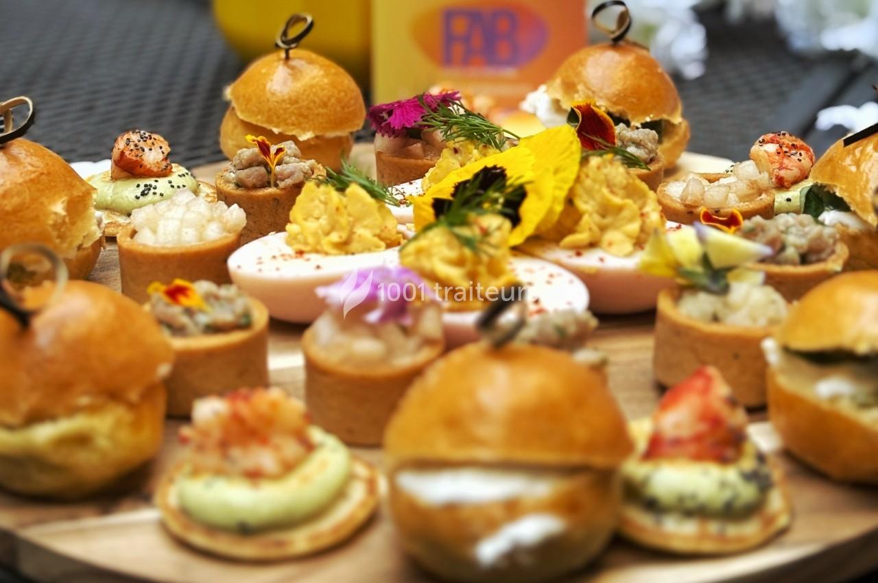 Plateau de bouchées apéritives variées comprenant mini-burgers, tartelettes, œufs farcis et décorations florales.