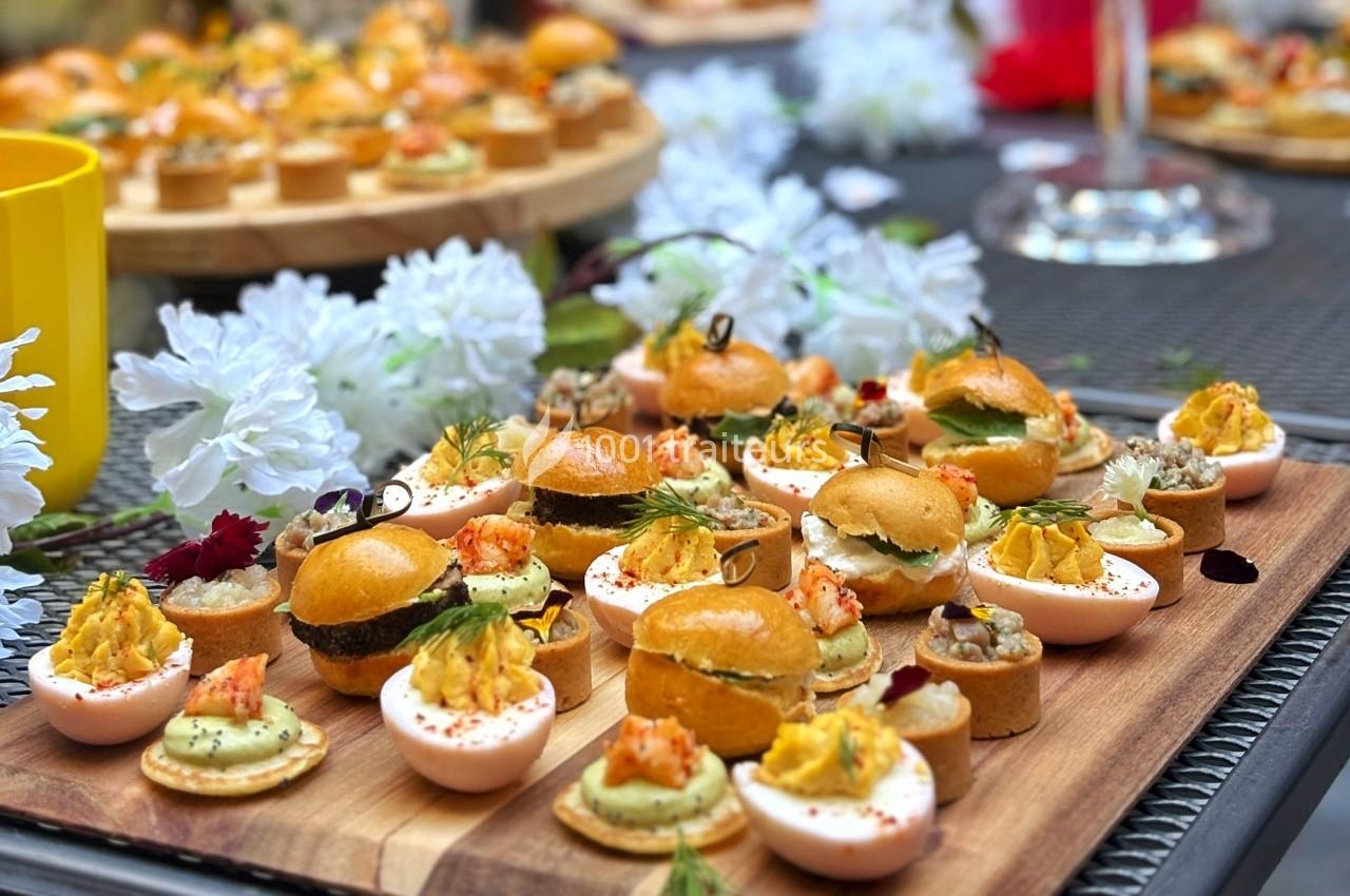 Plateau en bois garni de canapés variés, œufs farcis et mini-burgers, entouré de fleurs blanches décoratives.