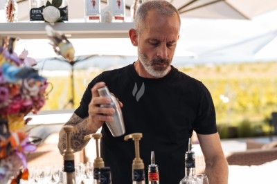 Un barman prépare un cocktail en utilisant un shaker derrière un comptoir extérieur, entouré de bouteilles et d'ombrelles.