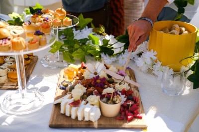 Plateau de fromages et apéritifs variés sur une table décorée de fleurs et de verdure, avec une main servant un plat.