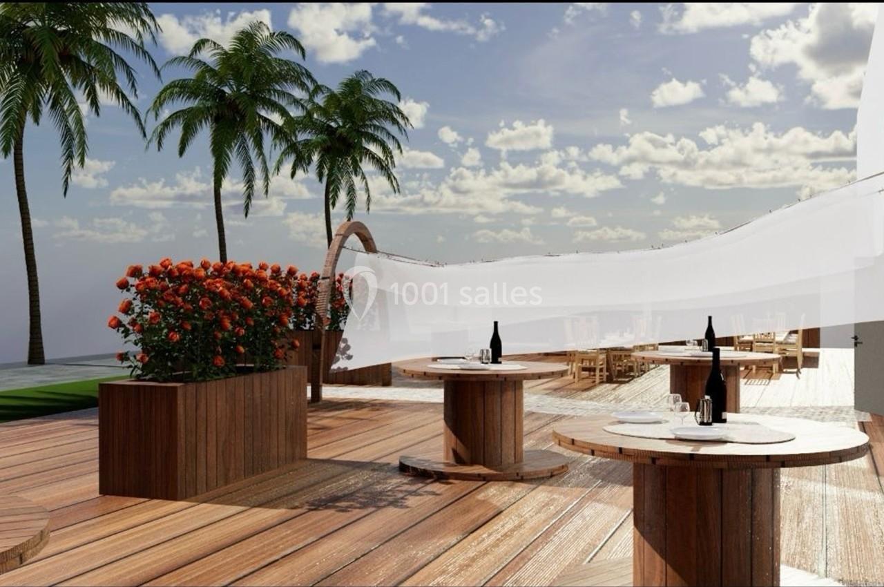 Terrasse en bois avec tables dressées, plantes fleuries et palmiers sous un ciel partiellement nuageux.