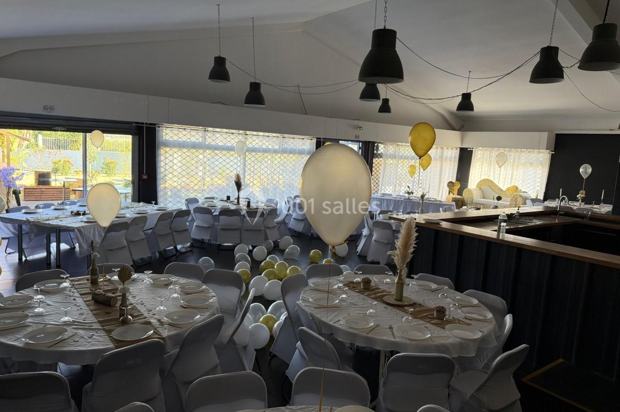 Salle décorée pour un événement, avec des tables rondes dressées, des ballons blancs et jaunes, et une lumière naturelle.