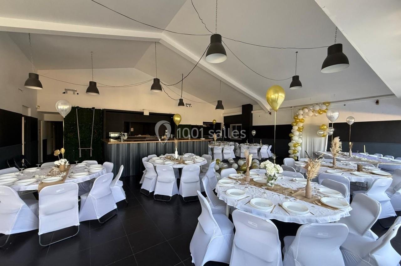 Salle de réception décorée avec des tables rondes, nappes blanches, ballons dorés et guirlandes lumineuses.