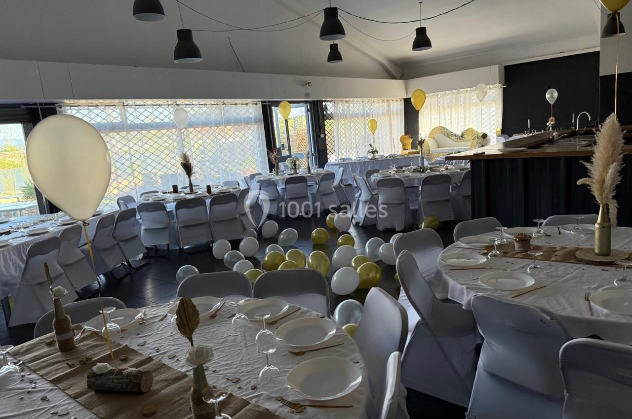 Salle décorée pour une réception avec tables dressées, ballons blancs et dorés au sol, et lumière naturelle.