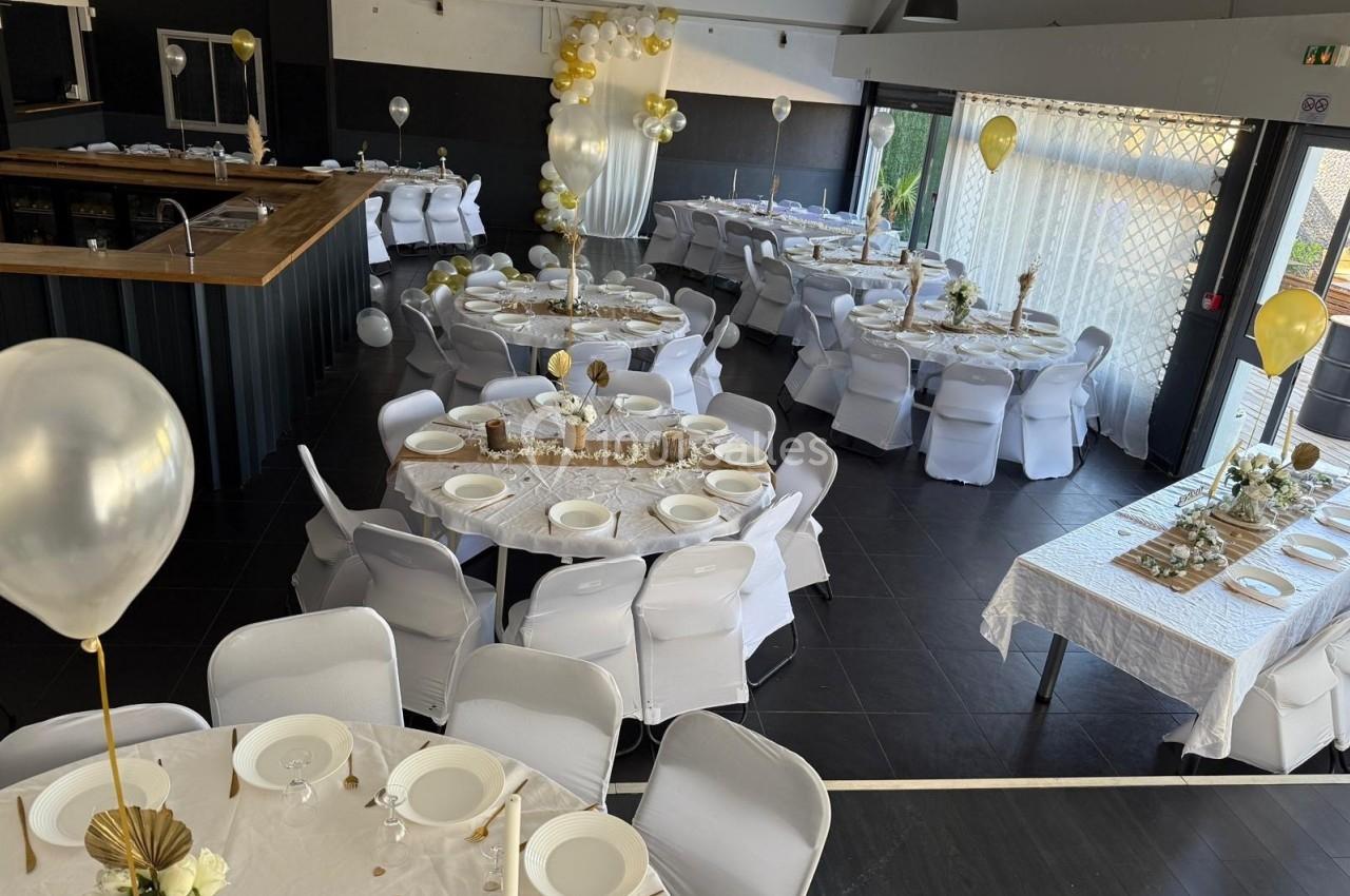 Salle décorée pour un événement avec des tables rondes et rectangulaires dressées, ornées de ballons et de nappes blanches.