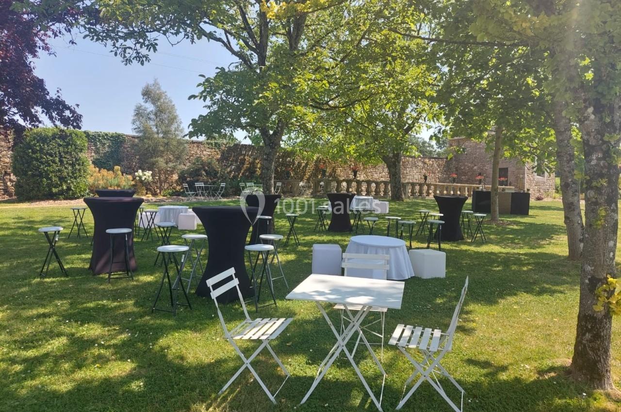 Tables et chaises blanches disposées sur une pelouse ombragée, entourées de tables hautes avec nappes noires.