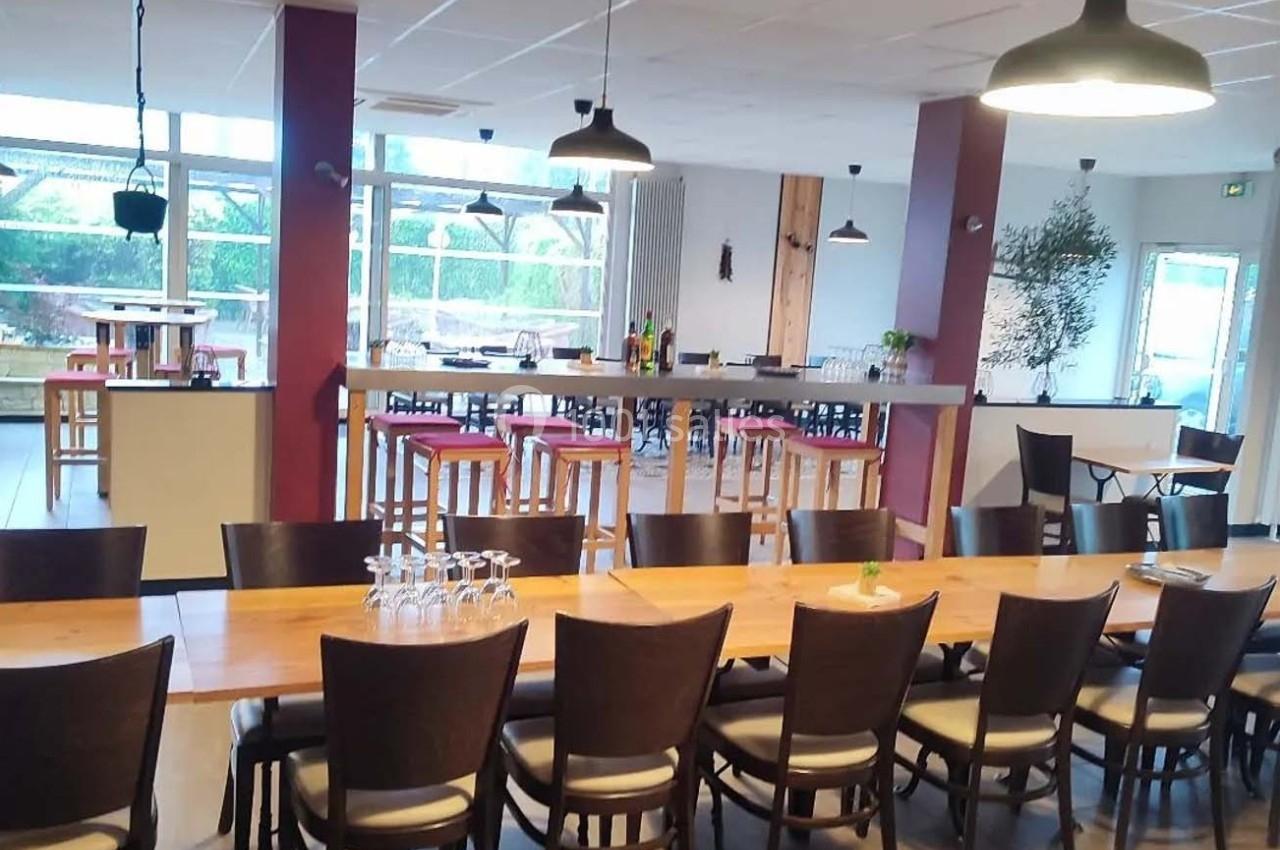 Salle de restaurant lumineuse avec grandes baies vitrées, tables en bois, chaises noires et suspensions modernes.