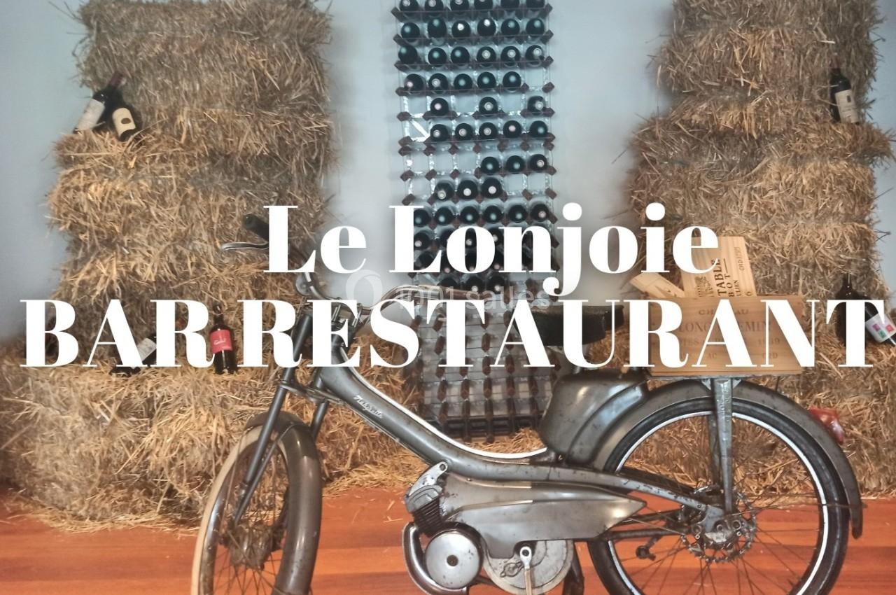 Une moto vintage devant des bottes de paille et un mur de bouteilles de vin alignées.