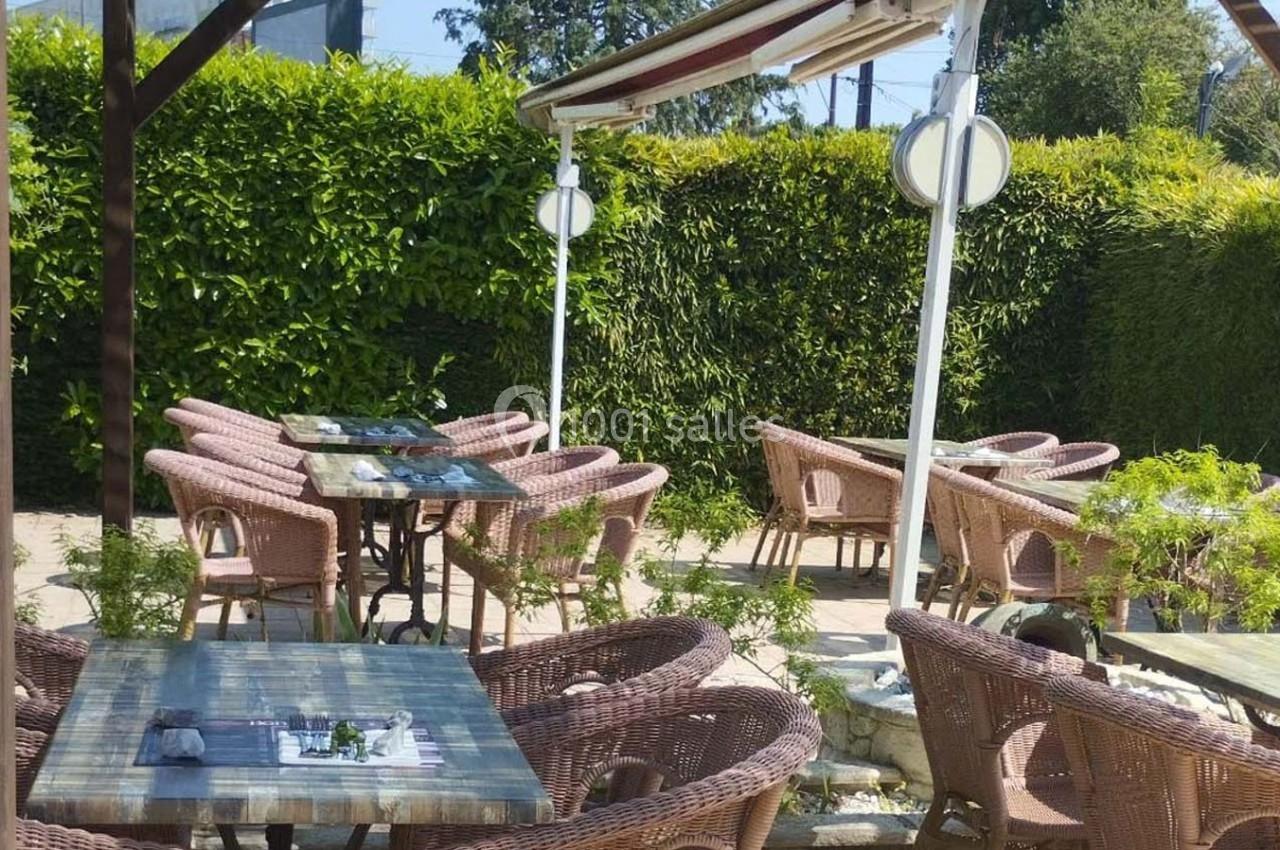 Terrasse extérieure avec tables en bois, chaises en rotin et végétation environnante sous un auvent.