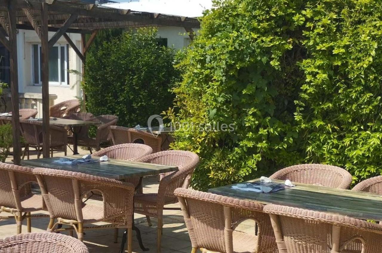 Terrasse extérieure avec tables et chaises en osier, entourée de verdure et partiellement ombragée par une pergola.
