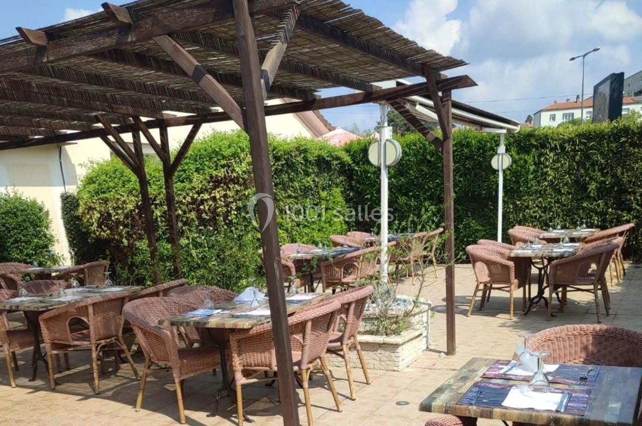 Terrasse extérieure avec tables et chaises en rotin sous une pergola en bois, entourée de haies verdoyantes.