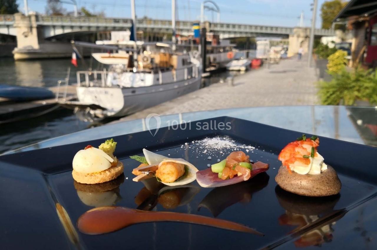 Assortiment de bouchées gastronomiques sur une assiette noire, avec un quai et des bateaux en arrière-plan.