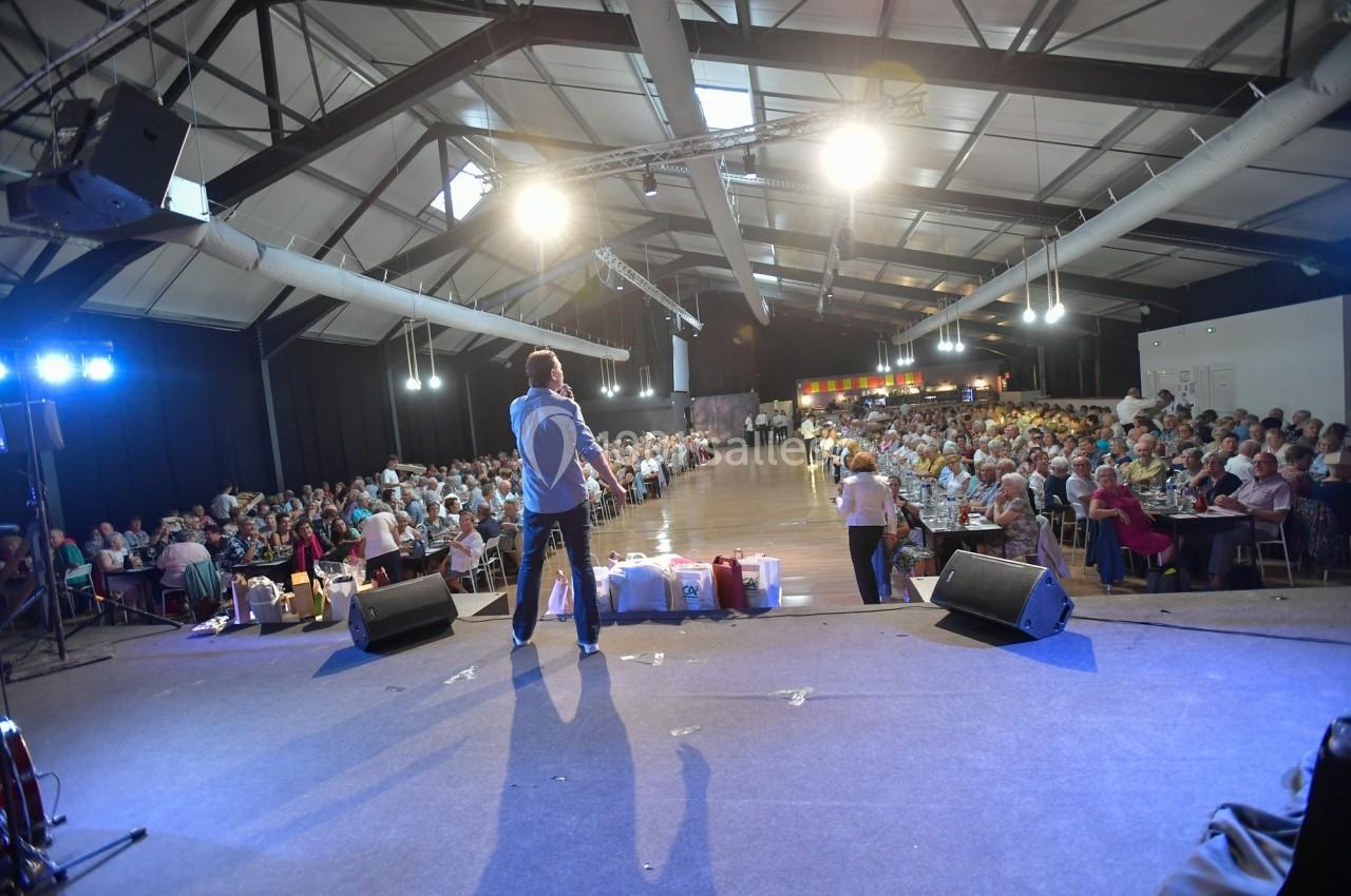 Un homme sur scène s'adresse à un large public assis dans une salle éclairée avec une structure métallique apparente.