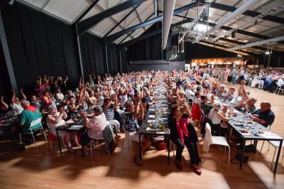 Un groupe de personnes photographiant un grand gâteau décoré, dans une salle remplie lors d'un événement festif.