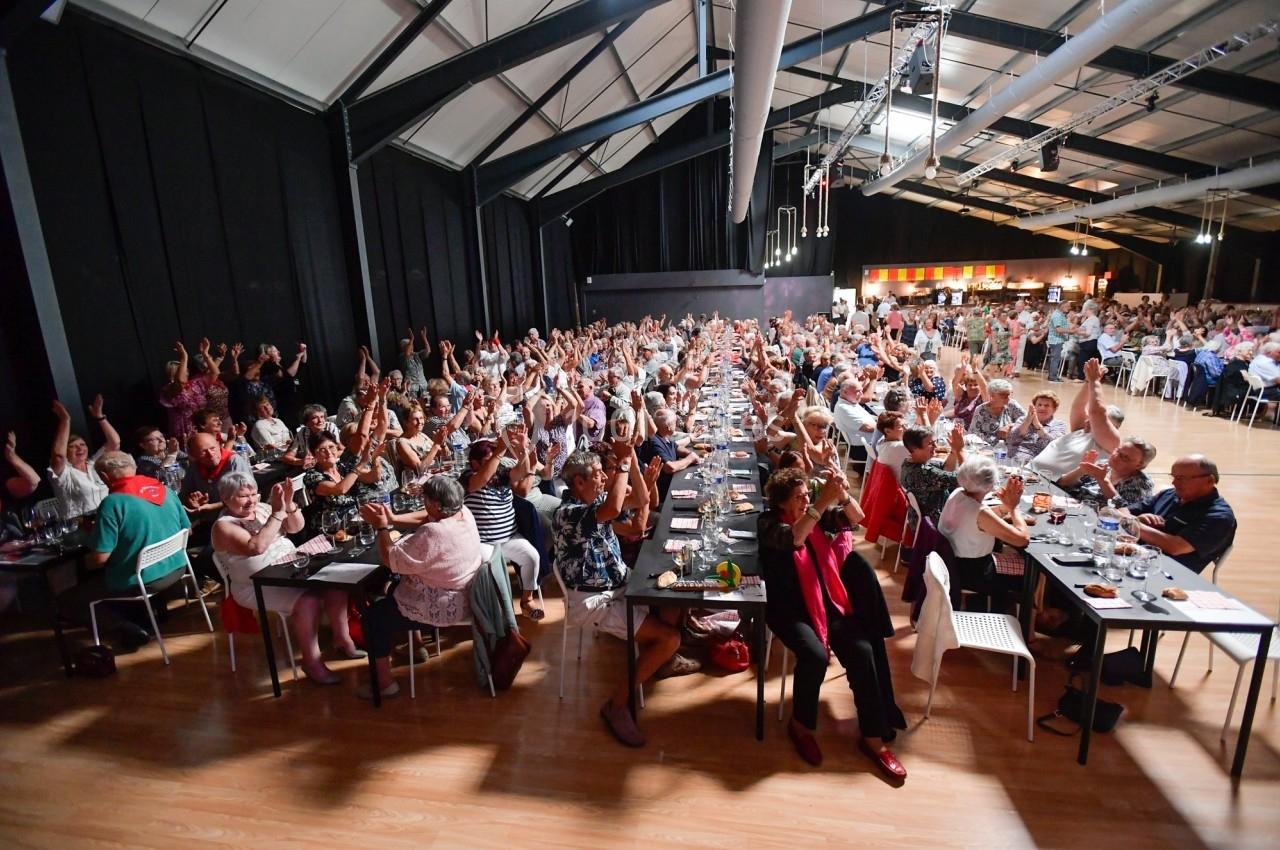 Des personnes assises à des tables dans une grande salle, levant la main lors d'un événement collectif.