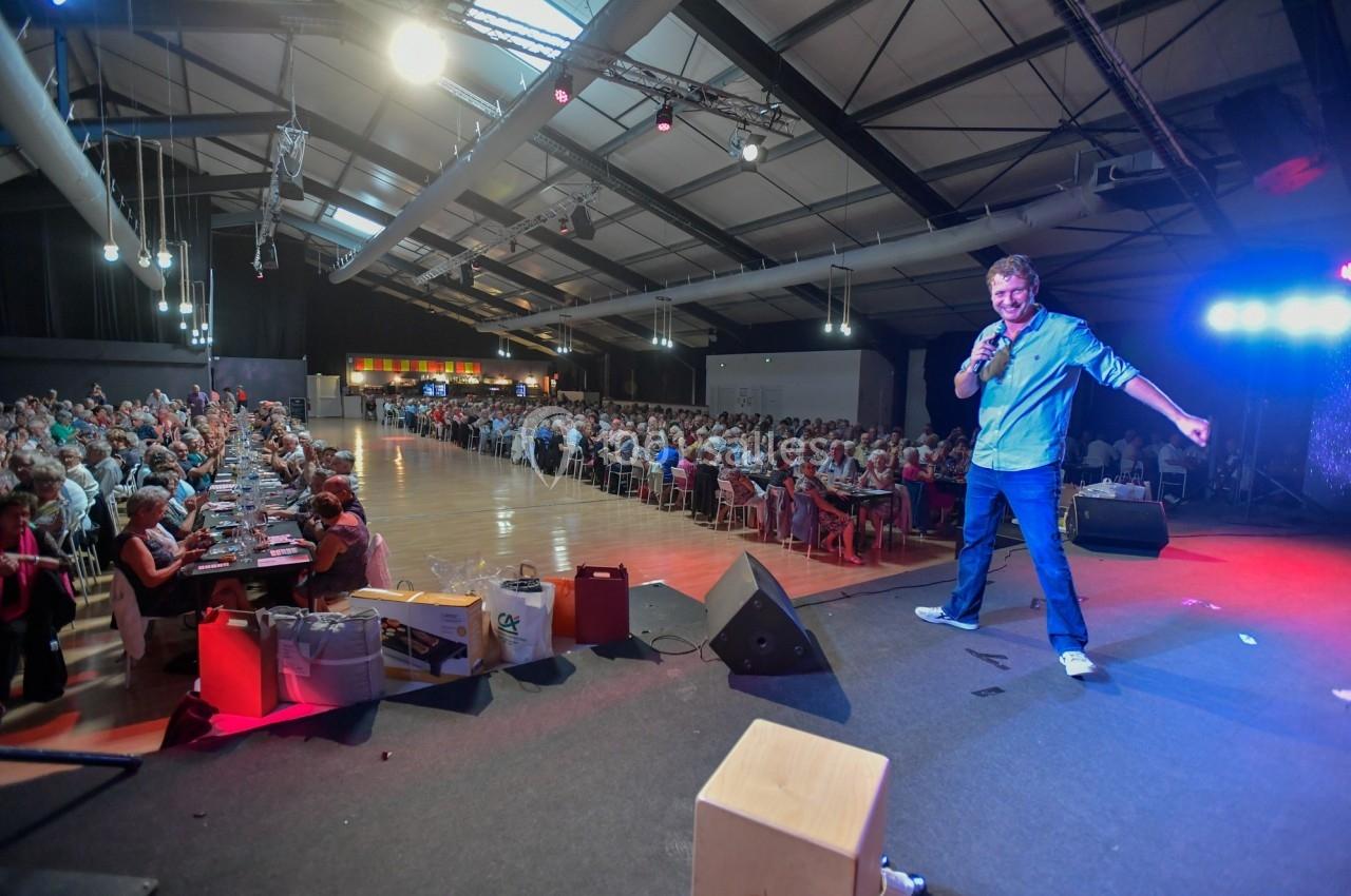 Un homme sur scène s'adresse à un large public assis dans une salle éclairée avec une ambiance conviviale.