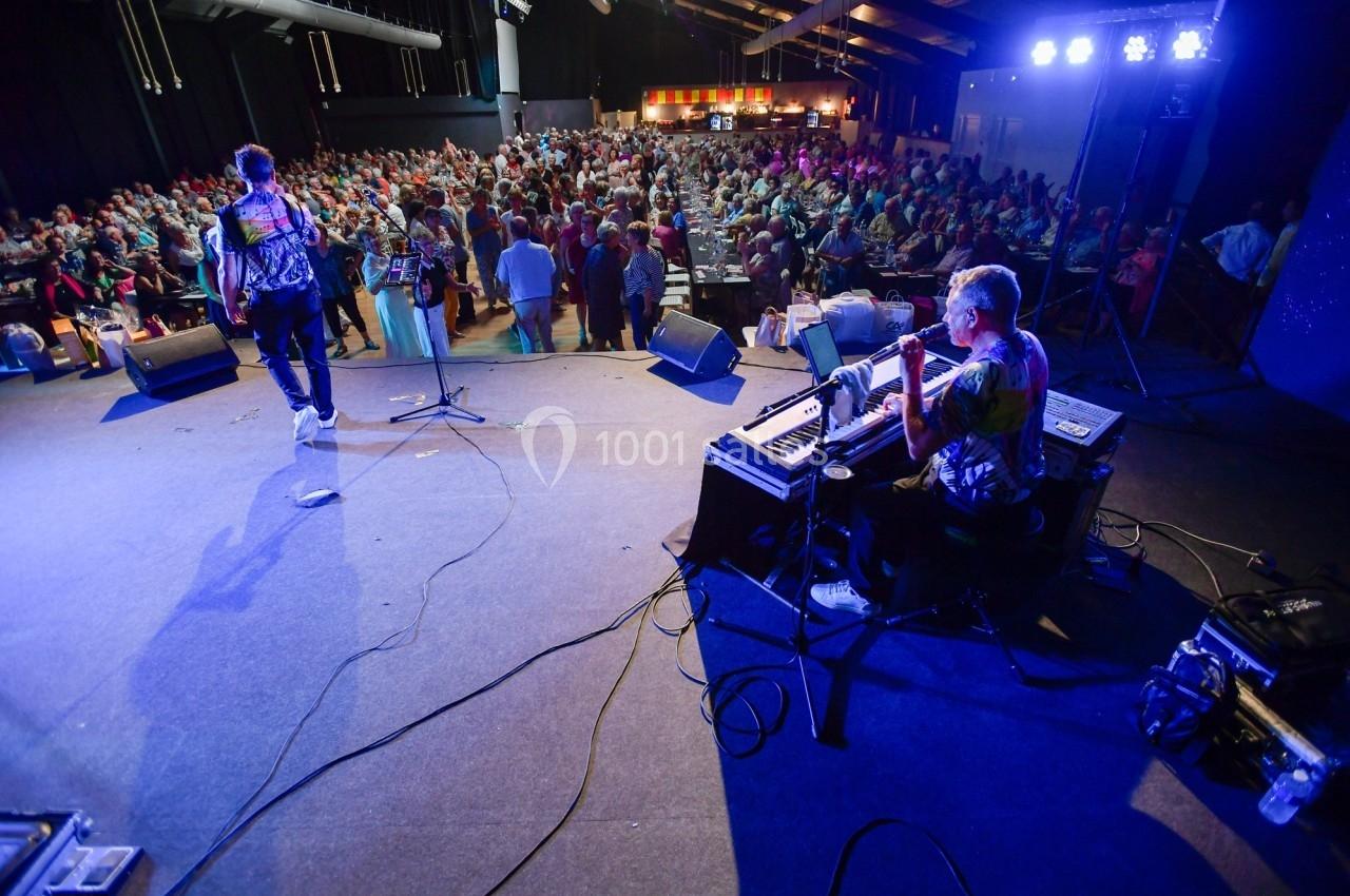 Vue depuis la scène d'un concert avec un musicien au clavier et une foule assise dans une salle éclairée.