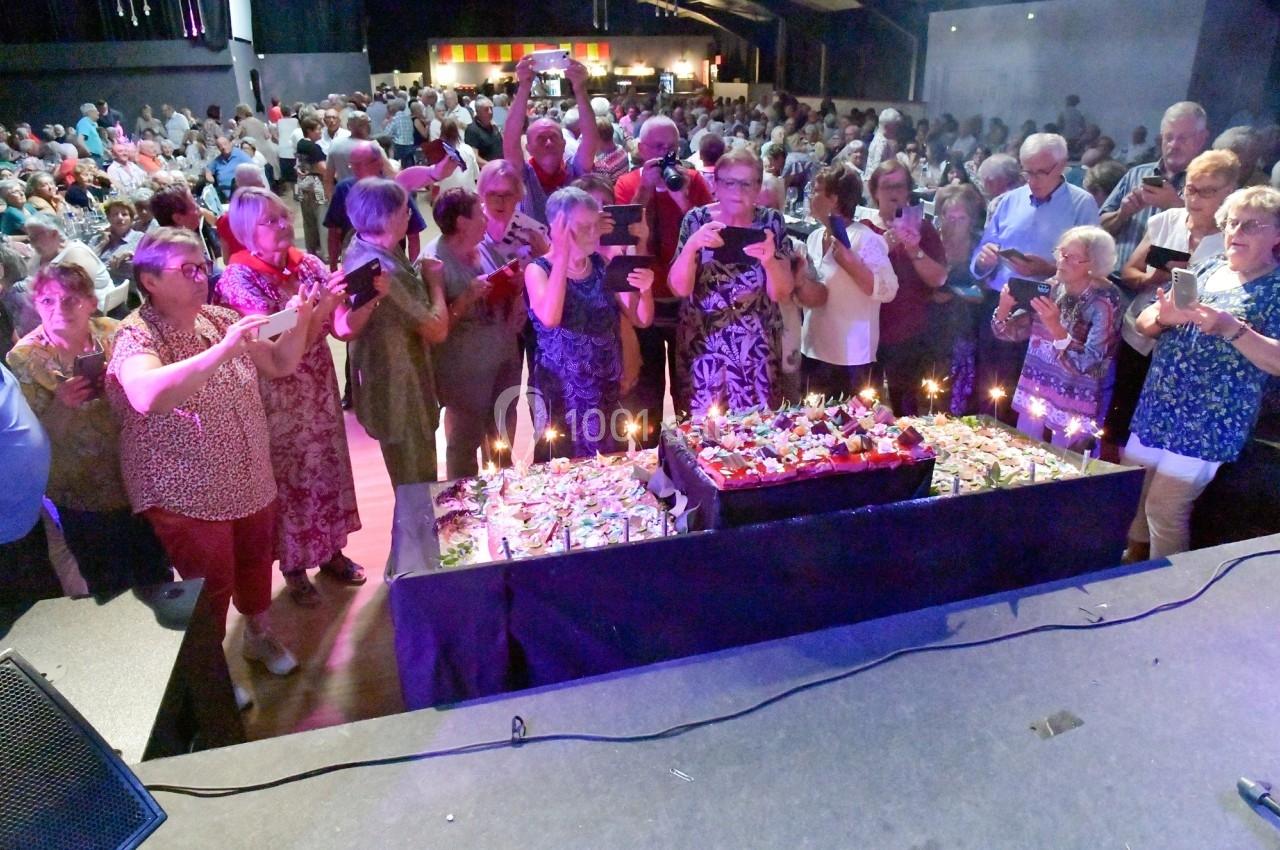 Un groupe de personnes photographiant un grand gâteau décoré, dans une salle remplie lors d'un événement festif.