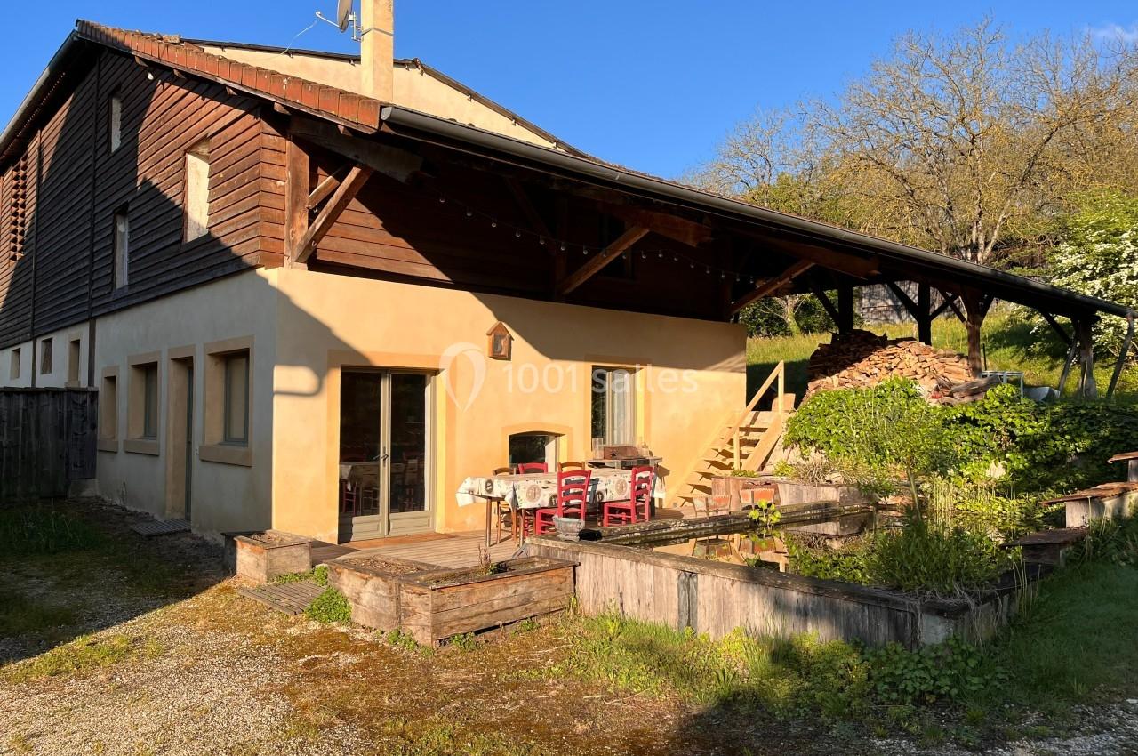 Maison en bois et crépi avec terrasse couverte, jardin aménagé et pile de bois à l'arrière, sous un ciel ensoleillé.