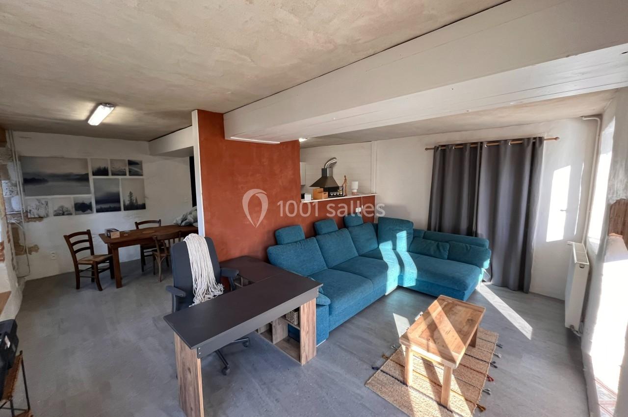 Salon lumineux avec canapé bleu, table basse en bois, bureau noir et espace salle à manger en arrière-plan.