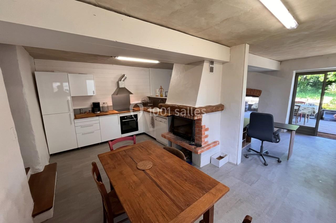 Cuisine ouverte avec table en bois, cheminée en briques, bureau et vue sur un espace extérieur verdoyant.