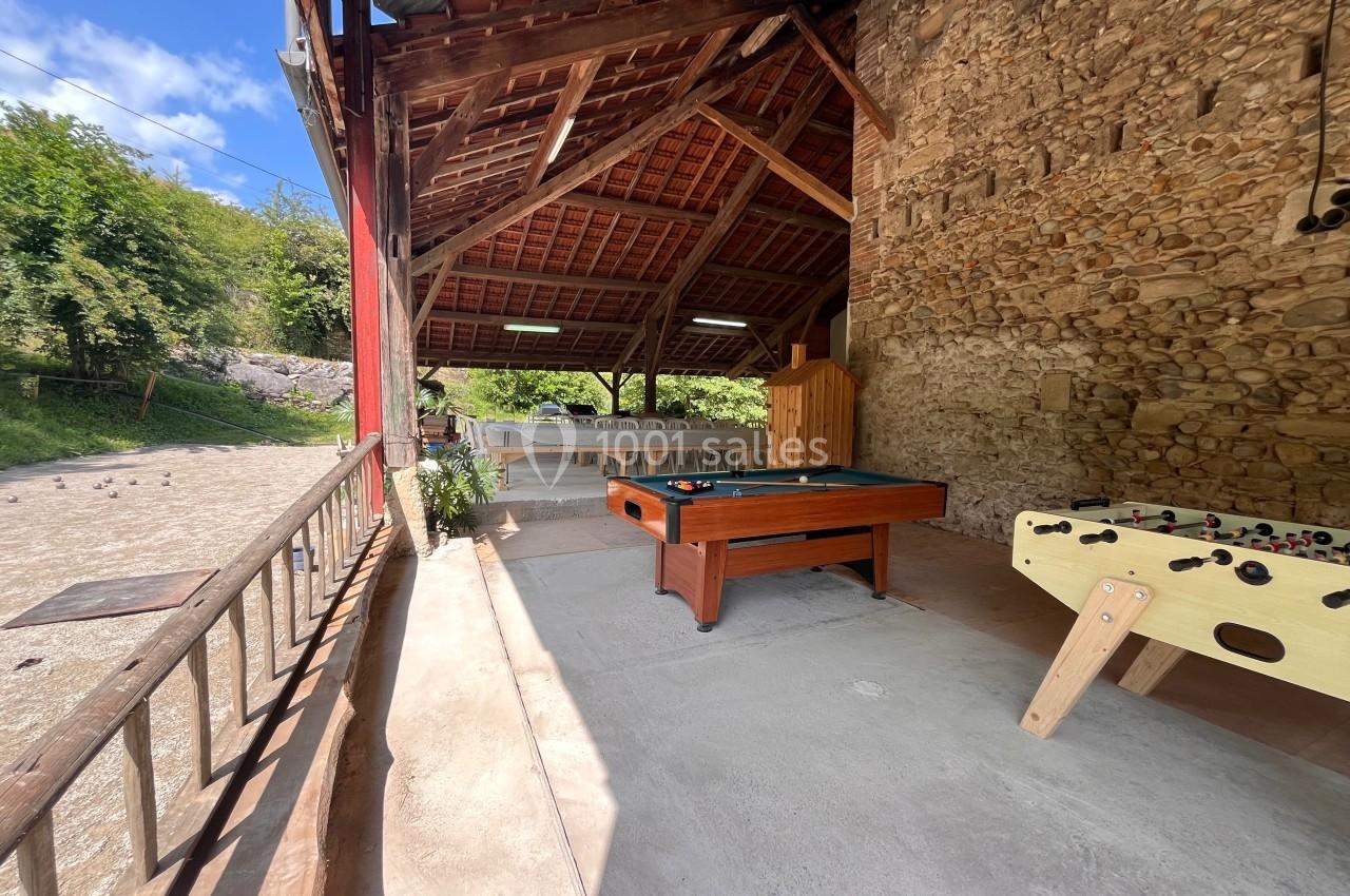 Espace couvert avec baby-foot, billard et tables, situé près d'un mur en pierre et ouvert sur l'extérieur verdoyant.