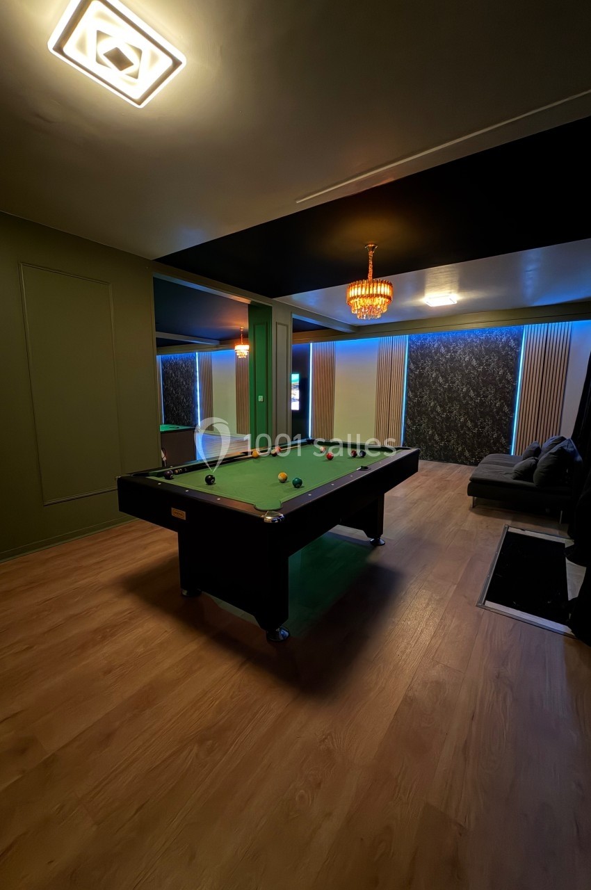 Salle avec un billard au centre, parquet au sol, éclairage tamisé et canapé dans un coin.
