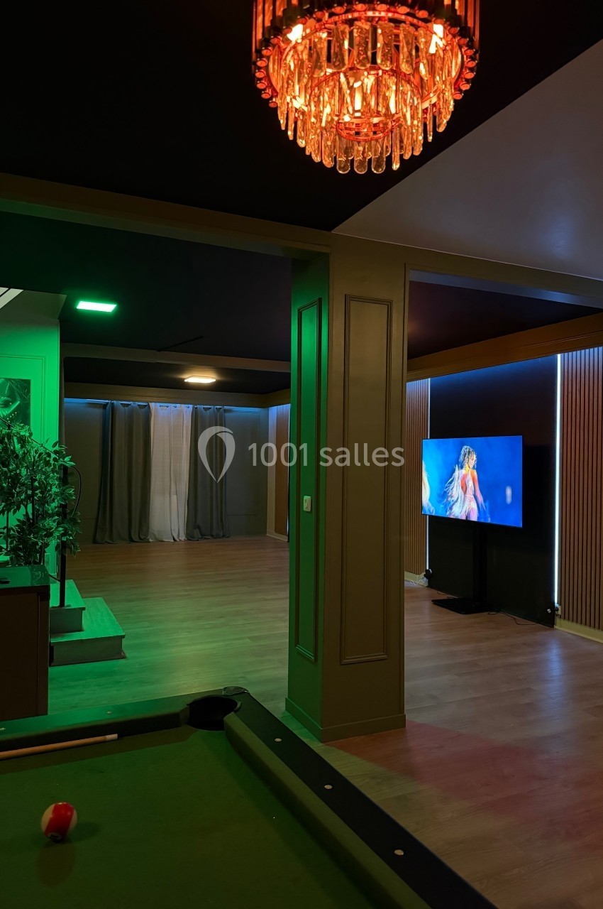 Salle de loisirs avec table de billard, télévision allumée et éclairage tamisé aux tons verts et orangés.