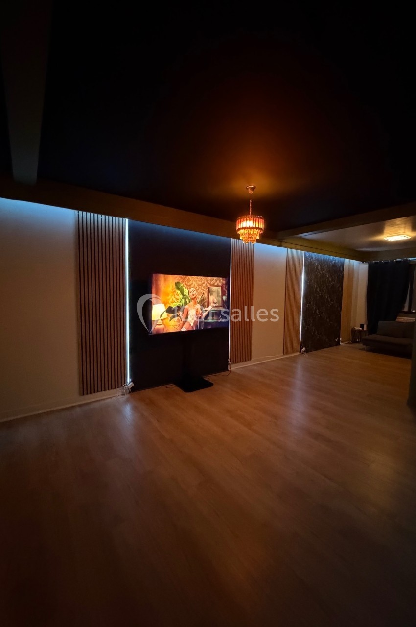 Salle sombre avec parquet, télé allumée affichant une image colorée, murs clairs et éclairage tamisé.