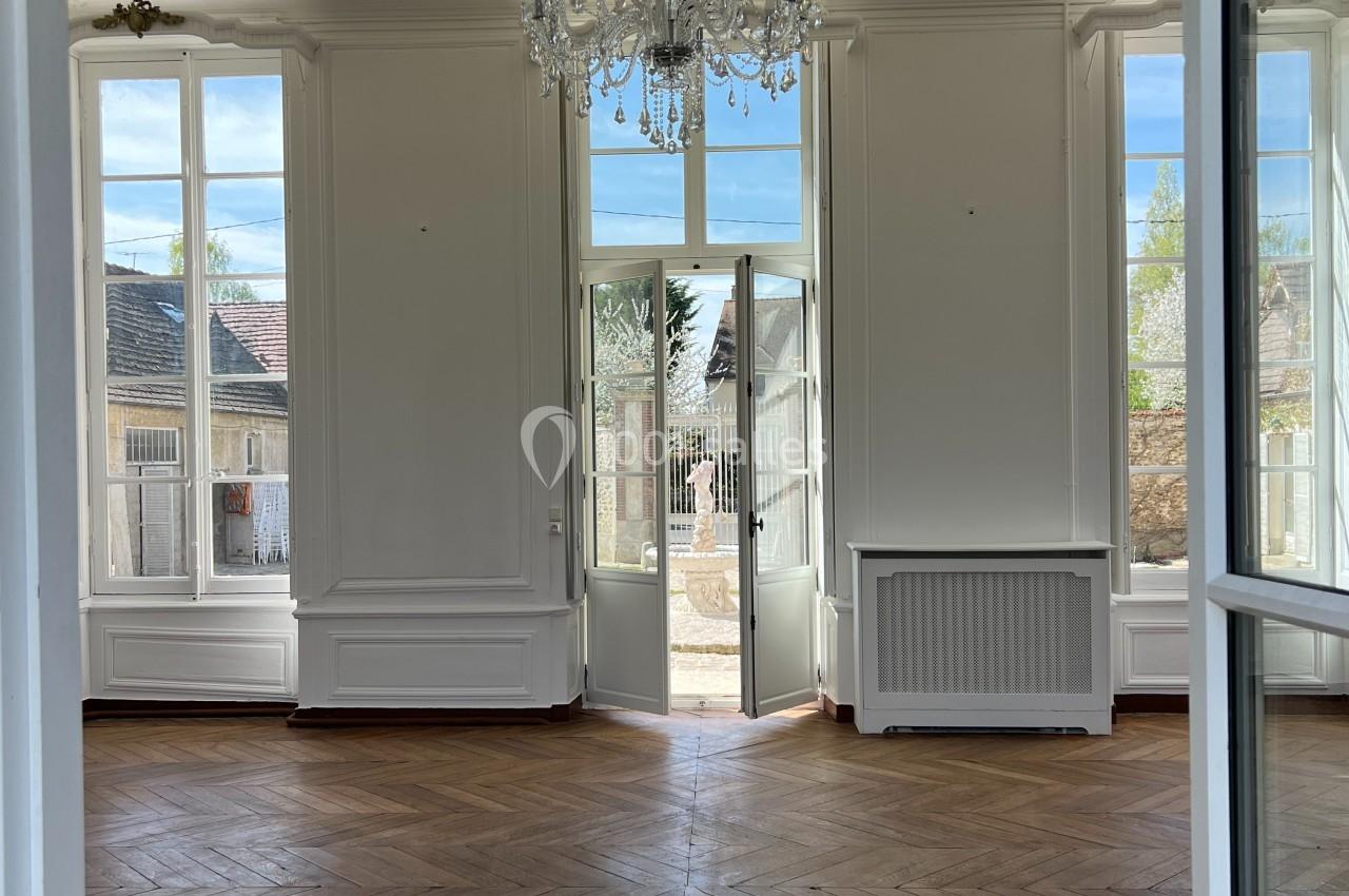 Salle lumineuse avec parquet en bois, grandes fenêtres, porte-fenêtre ouverte sur un jardin et lustre au plafond.