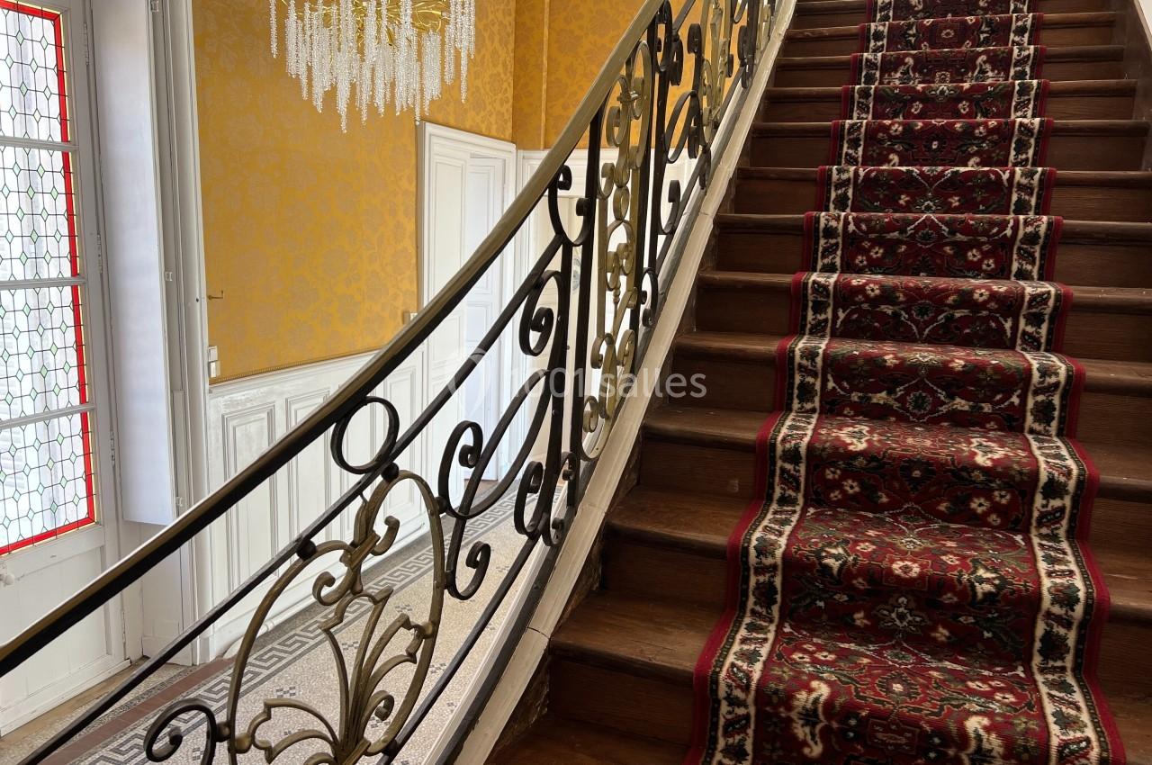 Escalier en bois avec rampe en fer forgé, tapis rouge à motifs et mur jaune orné d'un lustre en cristal.