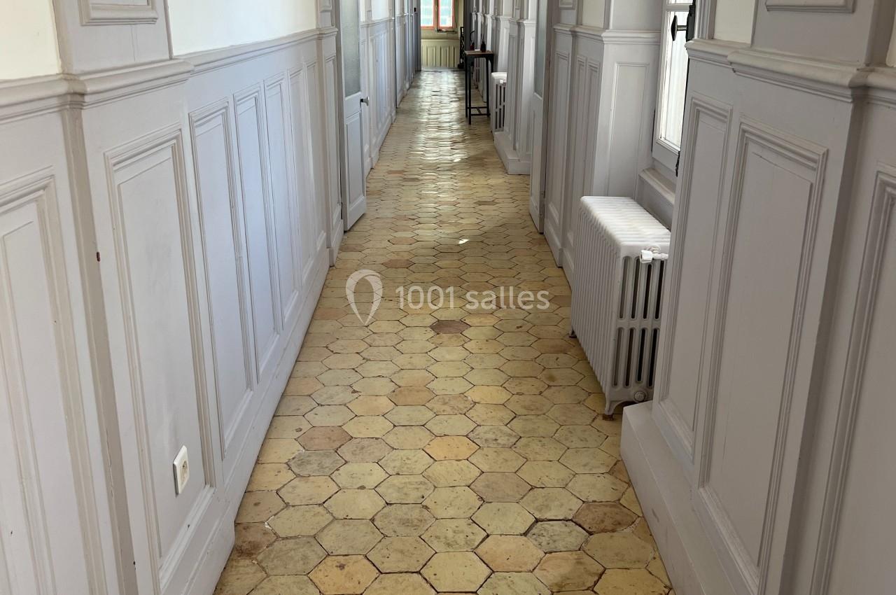 Couloir lumineux avec sol en carreaux hexagonaux, murs blancs et fenêtres laissant entrer la lumière naturelle.