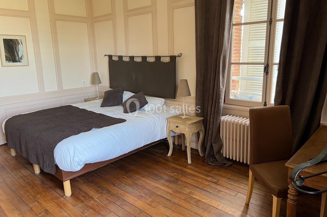 Chambre avec lit double, parquet en bois, table de chevet, chaise et fenêtre avec rideaux donnant sur l'extérieur.
